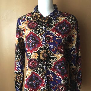 May include: A long-sleeved button-up shirt with a colorful geometric pattern in shades of red, blue, black, and gold. The pattern resembles a traditional rug or tapestry.