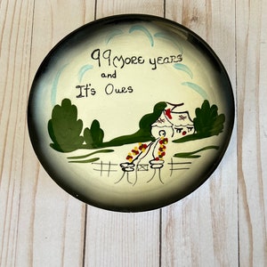 May include: A white ceramic plate with a black rim, featuring a hand-painted scene of a house with a red roof, green trees, and a fence. The plate has the text "99 more years and It's Ours" in black lettering.