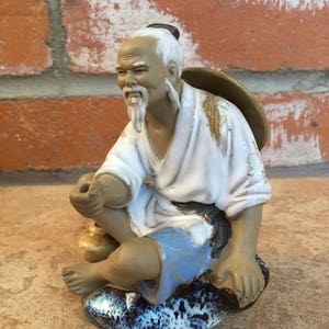 May include: A ceramic figurine of a man with a white beard and mustache, wearing a white robe and blue pants, sitting on a rock with a blue and white glaze. He is holding a small object in his hand.