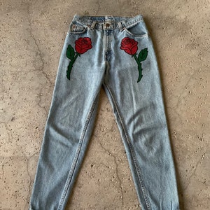 May include: Light blue denim jeans with two red roses embroidered on the front.