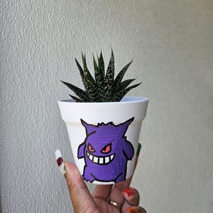 May include: A white ceramic plant pot featuring a purple cartoon character with red eyes and a wide grin. The pot holds a small succulent plant with dark green, pointed leaves. The pot is held in a hand with painted nails.
