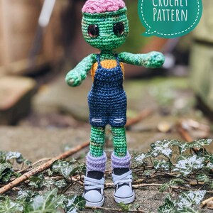 May include: A crocheted green zombie doll with a pink brain, blue overalls, and purple and white sneakers. The doll is standing on a bed of green leaves. The text "CROCHET PATTERN" is in a speech bubble above the doll.