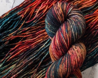 May include: A skein of hand-dyed yarn in a rich color palette of reds, oranges, and deep blues. The yarn is tightly wound, showcasing the vibrant color variations. The loose strands of yarn are spread out, displaying the texture and color transitions.