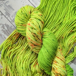 May include: A skein of vibrant green yarn with flecks of orange and yellow. The yarn is coiled, with loose strands extending outwards. The colour is a vivid, attention-grabbing shade of green, perfect for knitting or crochet projects.