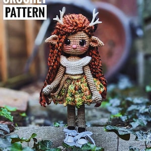 Op de afbeelding: Een gehaakte faunpop met bruin en oranje haar, die een wit topje en een groen, oranje en bruin rokje draagt. De pop heeft grote bruine ogen en draagt witte sneakers. De pop staat op een stenen oppervlak met groene bladeren.