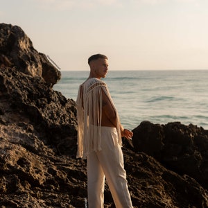 May include: A person wearing a white crocheted top with fringe and white pants stands on a rocky beach looking out at the ocean.