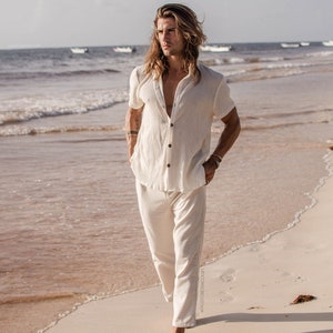 Puede incluir: Una persona con una camisa y pantalones de lino blanco camina por una playa de arena con el océano de fondo.