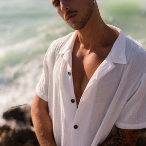 Puede incluir: Un hombre con una camisa blanca de manga corta abotonada de corte holgado. Tiene un tatuaje en el brazo y está de pie frente a una masa de agua.