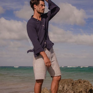 Puede incluir: Un hombre con una camisa de manga larga azul oscuro con cuello abotonado y pantalones cortos a rayas blancas. Está de pie en una playa rocosa con el océano de fondo. El cielo es azul con nubes blancas. El texto "#FAUSTOBETANCourt" es visible en la imagen.