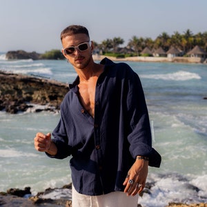 Puede incluir: Un hombre con una camisa azul marino de botones, pantalones blancos y gafas de sol blancas. Está de pie en una playa rocosa con el océano de fondo.