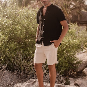 Puede incluir: Un hombre con una camisa negra de manga corta con botones y pantalones cortos de lino blancos está de pie sobre una roca. El hombre tiene el pelo largo y oscuro y está descalzo. El fondo es un entorno tropical con palmeras y vegetación exuberante. La imagen incluye el texto "#FAUSTOBETANCourt".