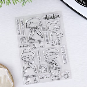 Op de afbeelding: Transparante stempels met illustraties van een meisje in een rode cape, een vrouw met een bril, een jongen met een hoed, een hond, een huis en een bol wol. De stempels bevatten Spaanse tekst, zoals "mi abuelita" en "dia de nietos".
