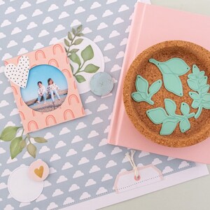 May include: A pink scrapbook page with a blue and white cloud pattern. The page features a photo of two children, a heart-shaped embellishment, and a green leaf design. A cork coaster with green leaf cutouts is also shown.