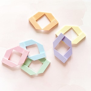 Puede incluir: Seis formas geométricas de plástico de color pastel. Las formas son todas hexágonos con un agujero cuadrado en el centro. Los colores son azul claro, verde claro, rosa claro, morado claro, amarillo claro y naranja.