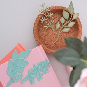 May include: Two die-cut paper floral designs in a light green color. The designs are placed on a cork coaster and a pink notebook.