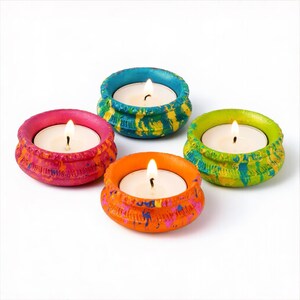 May include: Four colorful candle holders with lit candles. The holders are painted in bright hues of pink, blue, orange, and green, with textured designs. Each candle holder contains a white candle with a burning flame.