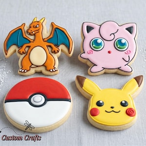 May include: Four decorated sugar cookies featuring popular characters. Includes a Charizard with orange and blue details, a pink Jigglypuff, a red and white Poké Ball, and a yellow Pikachu with red accents. The cookies are on a light gray surface.