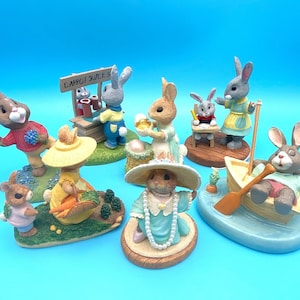 May include: A set of nine hand-painted ceramic figurines of bunnies in various poses. The bunnies are dressed in different outfits and are holding various props, including a carrot, a book, and a boat. The figurines are all standing on a variety of bases, including a wooden stand, a boat, and a grassy patch. The figurines are all painted in bright colors and have a whimsical and charming look.