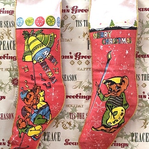 May include: Two red Christmas stockings with gold trim and white tops. One stocking features elves with musical notes and bells, the other has a cartoon character holding a paintbrush. Both say "Merry Christmas".