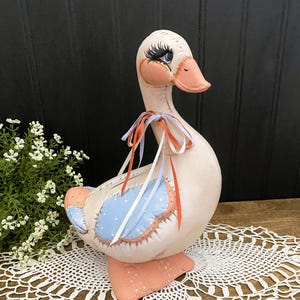 Könnte beinhalten: Eine ausgestopfte Stoffgans-Figur mit cremefarbenem Körper, orangefarbenem Schnabel und Füßen mit weißen Punkten sowie blauen Flügelpartien mit weißen Punkten. Die Gans hat aufgemalte Augen mit langen schwarzen Wimpern und ein dünnes orange-blaues Band.