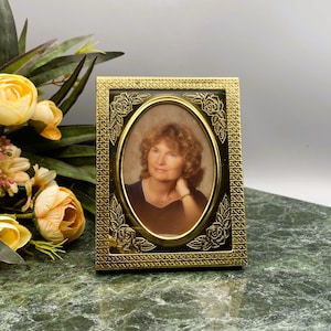 Puede incluir: Un marco de fotos dorado con un diseño adornado con rosas. El marco contiene una foto de una mujer con cabello castaño y una camisa blanca.
