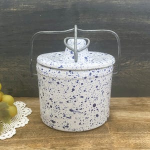 May include: A white ceramic jar with a lid and a metal handle. The jar is decorated with blue speckles. The lid has a small handle. The jar is sitting on a wooden surface.
