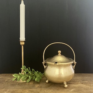 May include: A brass lidded pot with a swing handle and three ball feet, featuring a small notch in the rim for a spoon. Beside it stands a tall brass candlestick holding a white taper candle, accompanied by a small green leafy sprig.