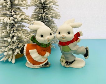 May include: Two ceramic bunny figurines. One bunny wears an orange and white dress with a green scarf, the other wears a gray outfit and red scarf, both on ice skates. Decorative winter holiday theme.