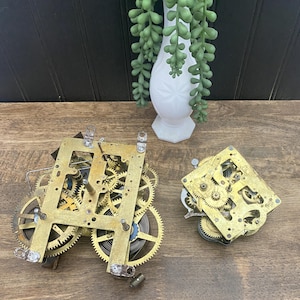 May include: Two brass clock movements featuring exposed gear trains, metal plates, and coiled springs. The larger mechanism is rectangular with multiple interlocking gears, while the smaller square unit displays a complex arrangement of brass cogs.
