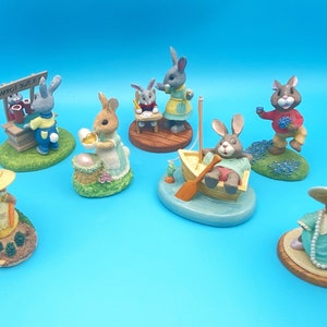 Puede incluir: Una colecci&oacute;n de siete figuritas de cer&aacute;mica pintadas a mano de conejos en varias poses. Los conejos llevan ropa y sostienen objetos, como una cesta de huevos, un puesto de zumo de zanahoria, un libro y un barco. Las figuritas est&aacute;n todas sobre peque&ntilde;as bases con hierba verde y flores.