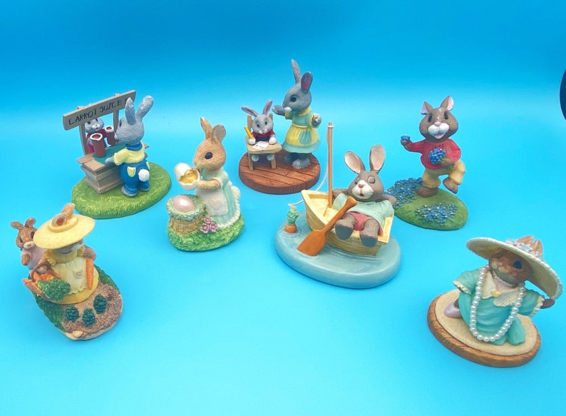 Puede incluir: Una colecci&oacute;n de siete figuritas de cer&aacute;mica pintadas a mano de conejos en varias poses. Los conejos llevan ropa y sostienen objetos, como una cesta de huevos, un puesto de zumo de zanahoria, un libro y un barco. Las figuritas est&aacute;n todas sobre peque&ntilde;as bases con hierba verde y flores.