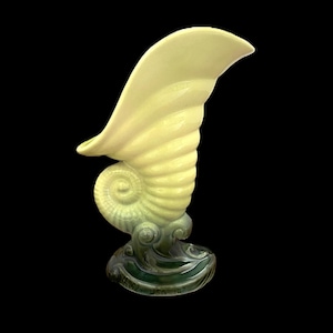 May include: A ceramic seashell-shaped vase in shades of yellow and green. The vase has a spiraled body and a curved, wing-like top. The base is dark green with wave-like details. A decorative piece for home decor.