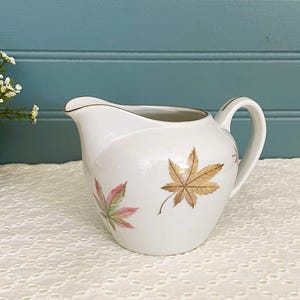 May include: White ceramic creamer with a gold rim and handle. The creamer is decorated with autumn-colored maple leaf designs in shades of brown, pink, and green. The creamer is a classic shape, perfect for serving cream or milk.