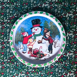 Puede incluir: Una placa de hojalata vintage con un muñeco de nieve y tres niños. El muñeco de nieve lleva un sombrero de copa negro y una bufanda roja. Los niños llevan ropa de invierno y sostienen una escoba y un bastón de caramelo. La placa de hojalata tiene un borde verde y blanco con las palabras "paz", "amor" y "alegría" impresas alrededor del borde.