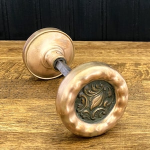 May include: A brass door knob set featuring a round, scalloped edge design with a dark, recessed center panel displaying an embossed floral pattern. The two knobs are connected by a square metal spindle, showcasing a metallic finish.