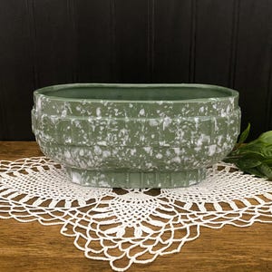May include: A green and white speckled ceramic planter with a rectangular shape. The planter has a textured, geometric design around the body and sits on a pedestal base. The planter is displayed on a white lace doily.