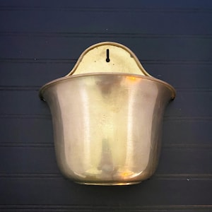 May include: A polished brass wall-mounted planter featuring a wide, curved opening and a flat back plate with a single vertical keyhole slot for hanging. The metallic surface has a warm, reflective golden tone with a smooth, rounded base.