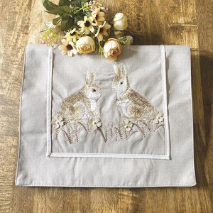 May include: A rectangular, light gray table runner with an embroidered design of two rabbits facing each other. The rabbits are brown and white, surrounded by embroidered flowers and grass. A white border frames the design.