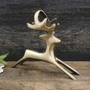 Puede incluir: Una escultura de reno estilizada de color latón. La escultura presenta un cuerpo esbelto, patas extendidas y astas prominentes. El reno está representado en posición de carrera, con la cabeza y las astas inclinadas hacia arriba.