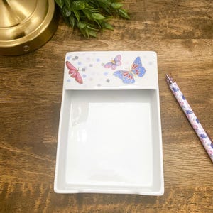 Puede incluir: Un plato rectangular de cerámica blanca con un diseño de mariposas. El plato tiene un borde elevado con tres mariposas coloridas y pequeños acentos cuadrados. Un bolígrafo estampado descansa sobre una superficie de madera.