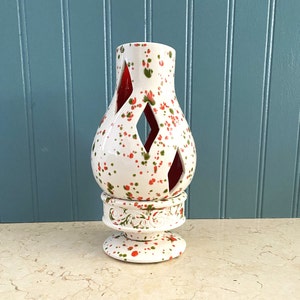 May include: White ceramic candle holder with red and green speckles. The holder has a cut-out design in the shape of diamonds. The base of the holder has a decorative design.