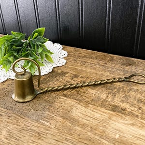 May include: A vintage brass candle snuffer with a bell-shaped head and a long, twisted handle. The snuffer is designed to extinguish candles safely. The handle has a loop at the end for hanging. The brass has a warm, aged patina.