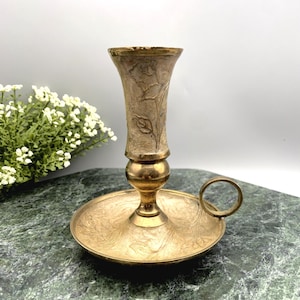 May include: A brass candlestick with a floral design. The candlestick has a round base with a handle and a tall, cylindrical holder.