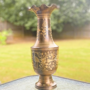May include: A vintage brass vase with a flared, ruffled top and a pedestal base. The vase is adorned with etched floral and bird designs. The overall color is a warm, antique gold, adding a touch of elegance to any space.