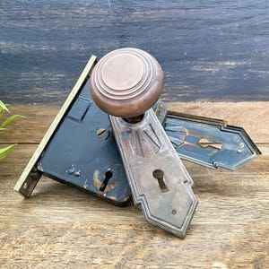 May include: Antique door hardware set featuring a bronze doorknob and decorative escutcheons. The set includes a black mortise lock with a brass strike plate. The hardware shows signs of age and wear, adding to its vintage appeal.
