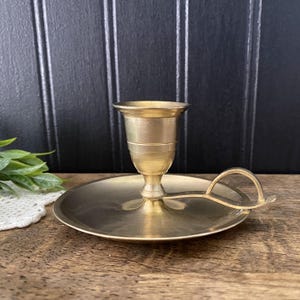 May include: A vintage brass candlestick holder with a round base and a handle. The candle holder has a cup-shaped top and a pedestal base. The brass has a warm, golden hue. The handle is a simple, curved design.