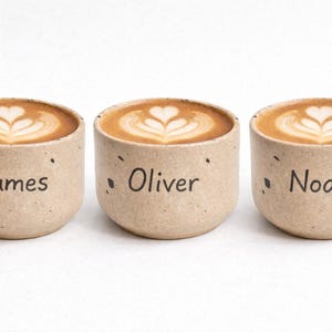 Op de afbeelding: Drie kleine, gespikkelde keramische bekers gevuld met koffie, elk met latte art. De bekers zijn voorzien van de namen "James", "Oliver" en "Noah" in zwarte tekst. De bekers zijn beige en staan tegen een witte achtergrond.