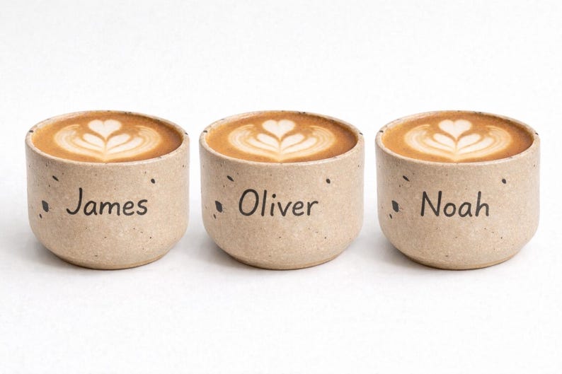 Op de afbeelding: Drie kleine, gespikkelde keramische bekers gevuld met koffie, elk met latte art. De bekers zijn voorzien van de namen "James", "Oliver" en "Noah" in zwarte tekst. De bekers zijn beige en staan tegen een witte achtergrond.