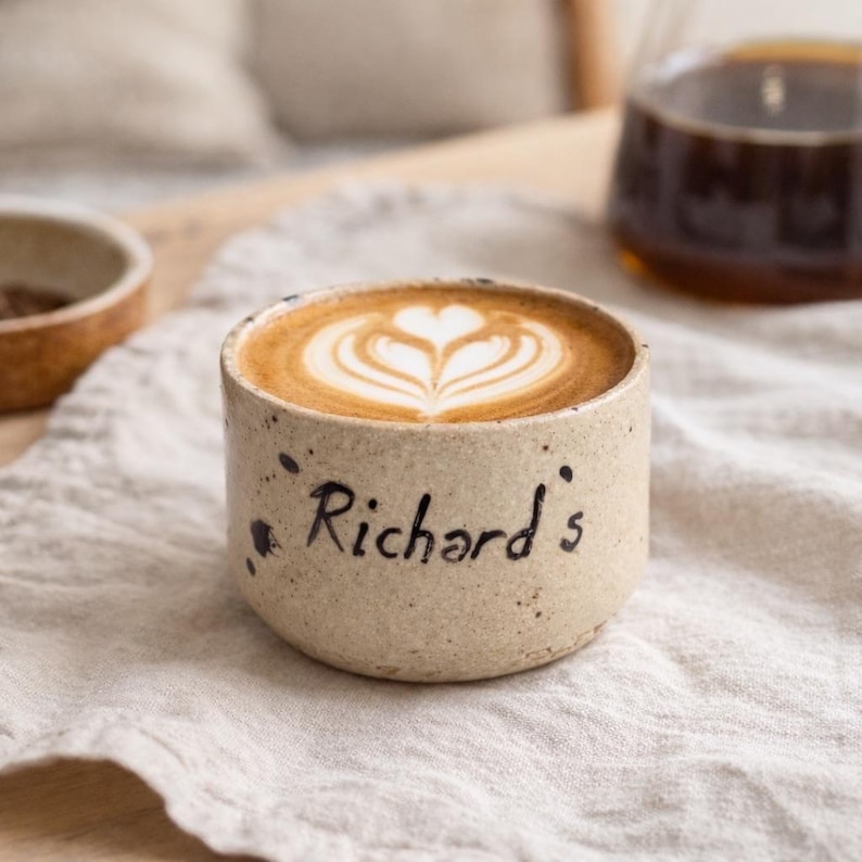 Op de afbeelding: Een beige keramische koffiekop met een latte art design. De beker heeft zwarte spikkels en de naam "Richard's" op de zijkant. De koffie heeft een hartvormig ontwerp. De beker staat op een witte doek.