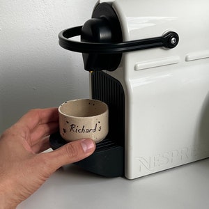Op de afbeelding: Een witte Nespresso-koffiemachine met zwarte details. Een kleine, gespikkelde keramische beker met de tekst "Richard's" staat op de lekbak van de machine. De machine staat op een wit oppervlak.